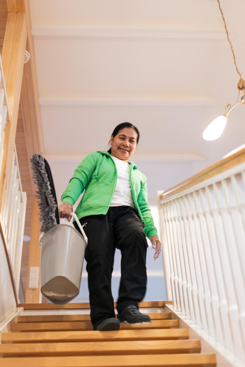 Emily Aguilar laskeutuu portaita hymyillen ja siivousvälineitä kantaen.